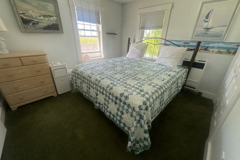 A small bedroom with a double bed covered in a green and white patterned quilt, a light wood dresser, and two windows with white blinds. A sailboat painting hangs on the wall above the bed.