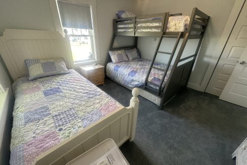 A bedroom with a white twin bed and a gray bunk bed with a twin over full configuration. Quilted bedspreads are on each bed, and a wooden nightstand is between the windows and the twin bed.