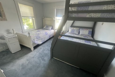 The photo shows a bedroom with one single bed and one bunk bed. The room has light-colored walls, carpeted floor, and two windows with blinds. An air conditioner stands to the left of the single bed.