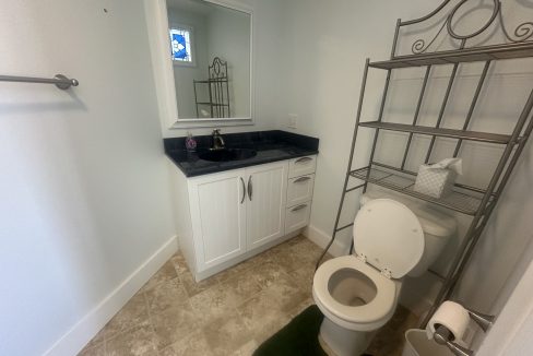 A small bathroom with a white vanity, a sink with a black countertop, a mirror, a metal shelf above the toilet, and a roll of toilet paper on a stand next to the toilet.