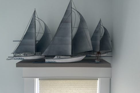 Three miniature sailboat models with grey sails are placed on a small shelf above a window with a beige pull-down shade.