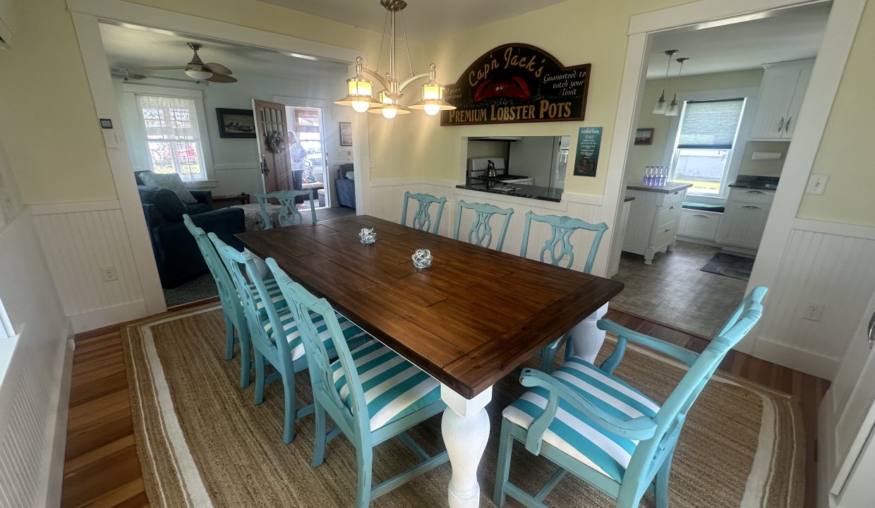 A dining room with a wooden table and eight blue and white striped chairs, a chandelier above, and an adjoining kitchen and living room in the background. A sign reads 
