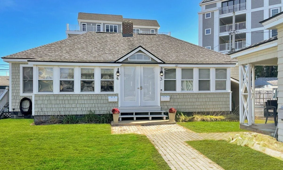 A small single-story white house with a gray shingled roof and central white door, flanked by two red planter boxes, with a stone walkway leading up to it on a sunny day.