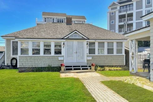 A small single-story white house with a gray shingled roof and central white door, flanked by two red planter boxes, with a stone walkway leading up to it on a sunny day.