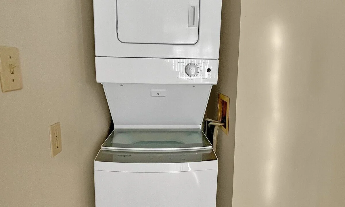 A stacked white washer and dryer set in a narrow hallway with wooden flooring and beige walls.