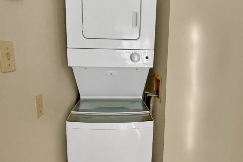 A stacked white washer and dryer set in a narrow hallway with wooden flooring and beige walls.