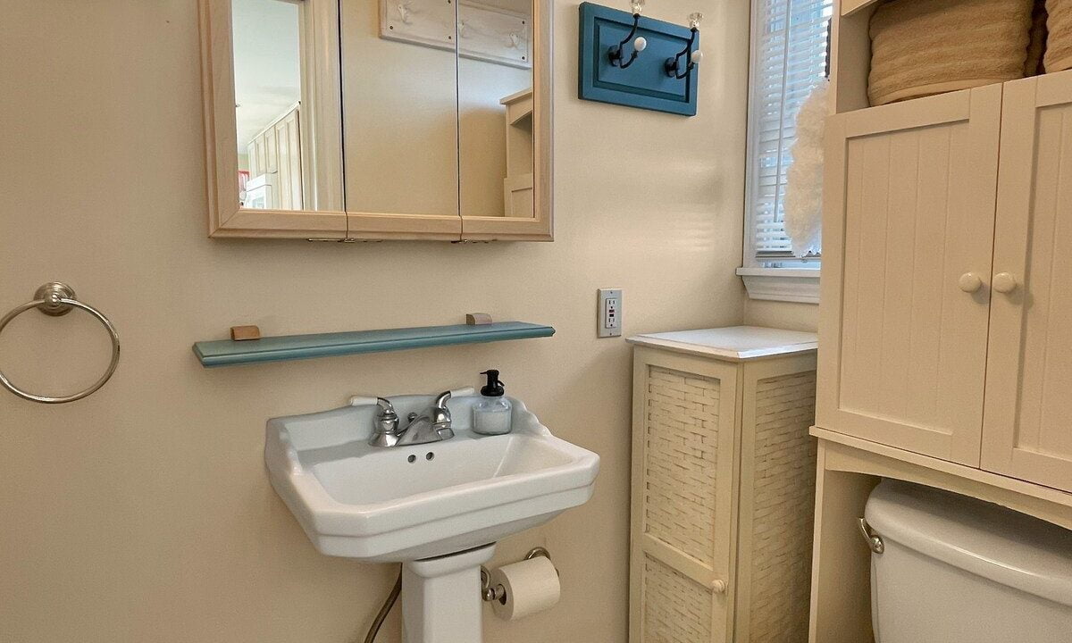 Interior of a small bathroom featuring a pedestal sink, toilet, and a mirrored medicine cabinet with rustic lighting above. light blue accents and wooden flooring are visible.