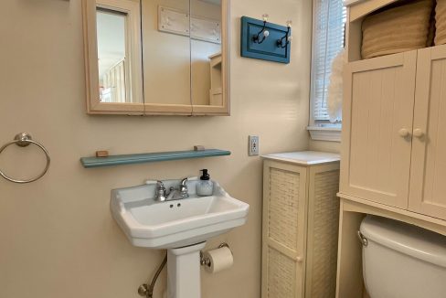 Interior of a small bathroom featuring a pedestal sink, toilet, and a mirrored medicine cabinet with rustic lighting above. light blue accents and wooden flooring are visible.