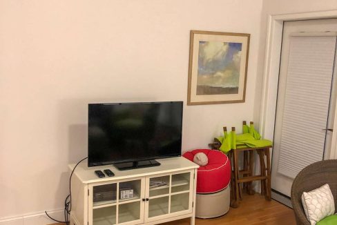 Living room corner with a wall-mounted air conditioner, a TV on a white console, children's toys, a framed painting, and a wicker chair.