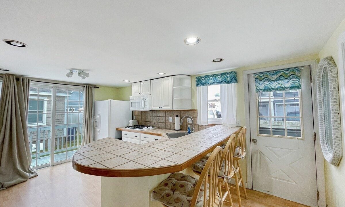 Bright kitchen with a curved island, white cabinets, and tile backsplash, leading to a balcony with draped curtains.