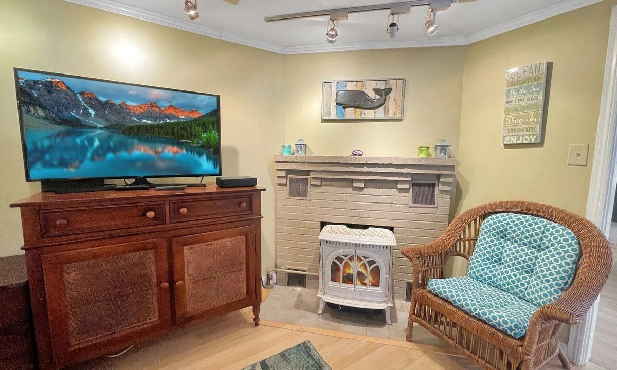 Cozy living room with a wicker chair, television on a wooden cabinet, fireplace, and decorative wall signs.