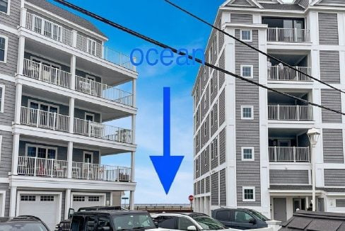 Three-story gray seaside condos flank a view of the ocean indicated by a blue arrow pointing downwards. The ocean is visible between the buildings. Parked cars are in the foreground.