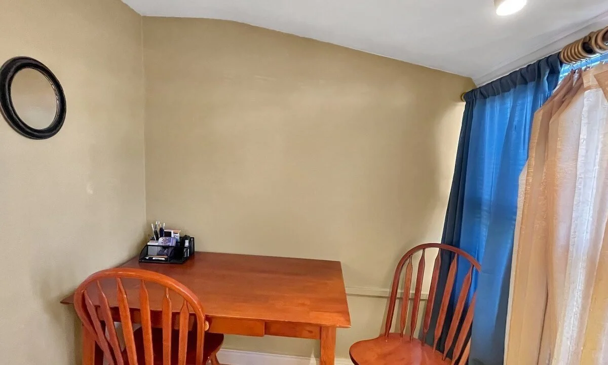 A small, cozy dining area with a wooden table and two chairs, near a window with blue and beige curtains and a mirror on the wall.