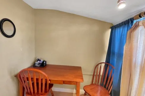 A small, cozy dining area with a wooden table and two chairs, near a window with blue and beige curtains and a mirror on the wall.