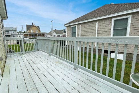 A spacious outdoor wooden deck with a white railing overlooks neighboring houses under a clear blue sky.