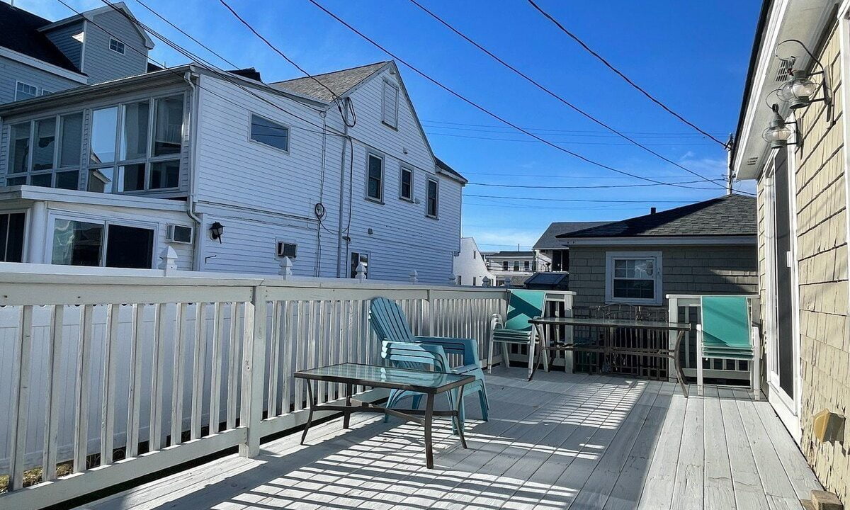 A sunny deck with outdoor furniture surrounded by white wooden fences and several residential buildings.