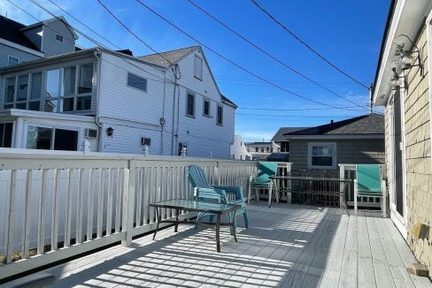 A sunny deck with outdoor furniture surrounded by white wooden fences and several residential buildings.