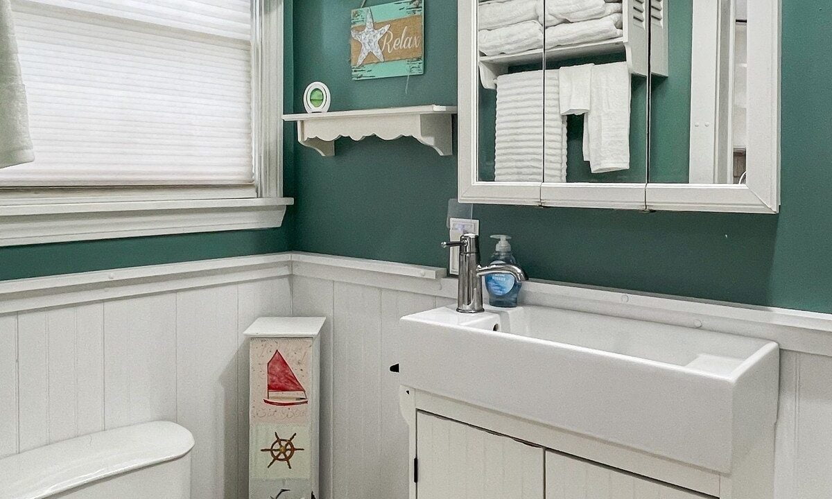 A small bathroom with teal walls, white wainscoting, and a cabinet sink. there is a toilet on the left and a mirror above the sink, with nautical decor elements.