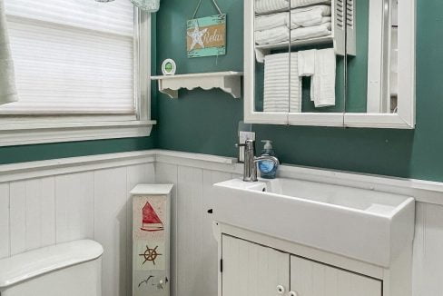 A small bathroom with teal walls, white wainscoting, and a cabinet sink. there is a toilet on the left and a mirror above the sink, with nautical decor elements.