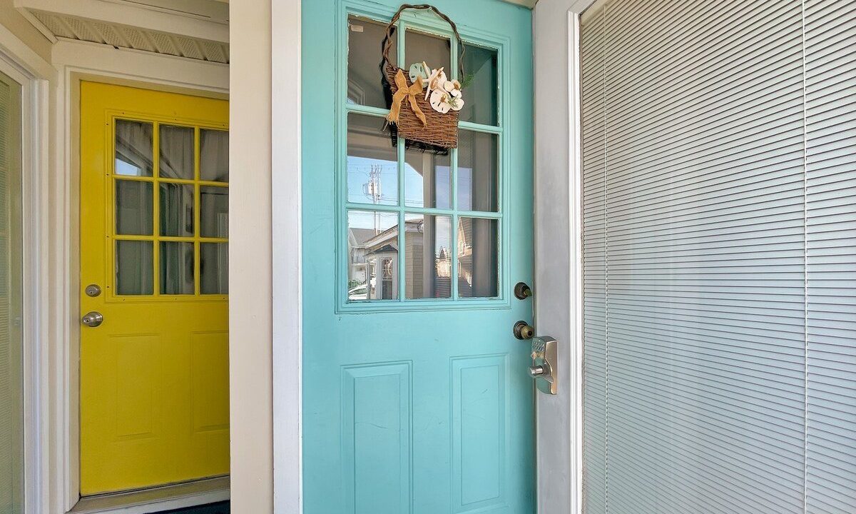 A turquoise door with a small hanging basket, partially open, revealing a yellow door across a bright hallway.