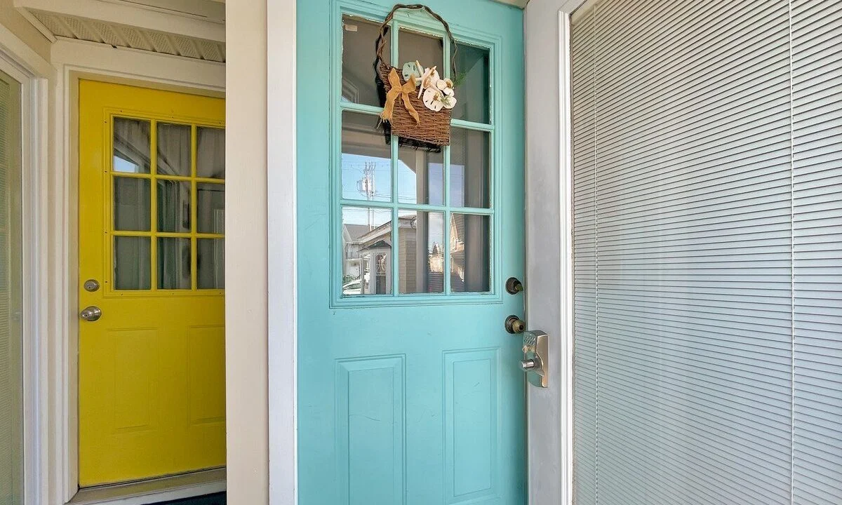 A turquoise door with a small hanging basket, partially open, revealing a yellow door across a bright hallway.