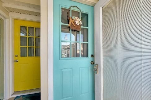 A turquoise door with a small hanging basket, partially open, revealing a yellow door across a bright hallway.