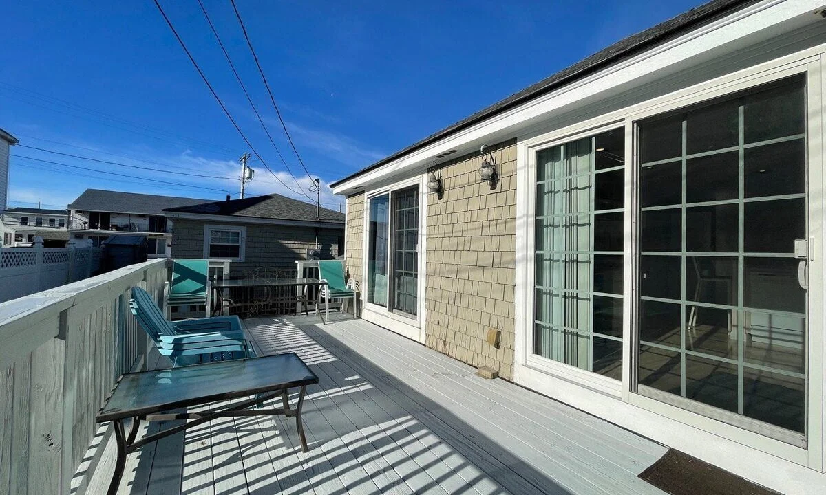 An exterior view of a house with a spacious wooden deck featuring a table, chairs, and a bench under a clear blue sky.