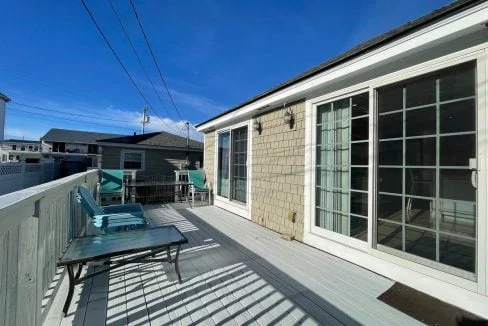 An exterior view of a house with a spacious wooden deck featuring a table, chairs, and a bench under a clear blue sky.