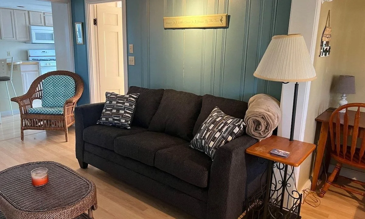 A cozy living room with a dark gray sofa, wicker chairs, wooden dining table, teal walls, and a kitchen visible in the background.