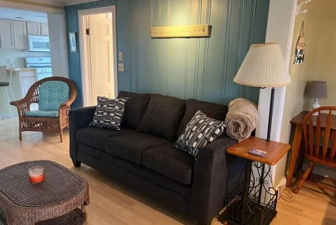 A cozy living room with a dark gray sofa, wicker chairs, wooden dining table, teal walls, and a kitchen visible in the background.