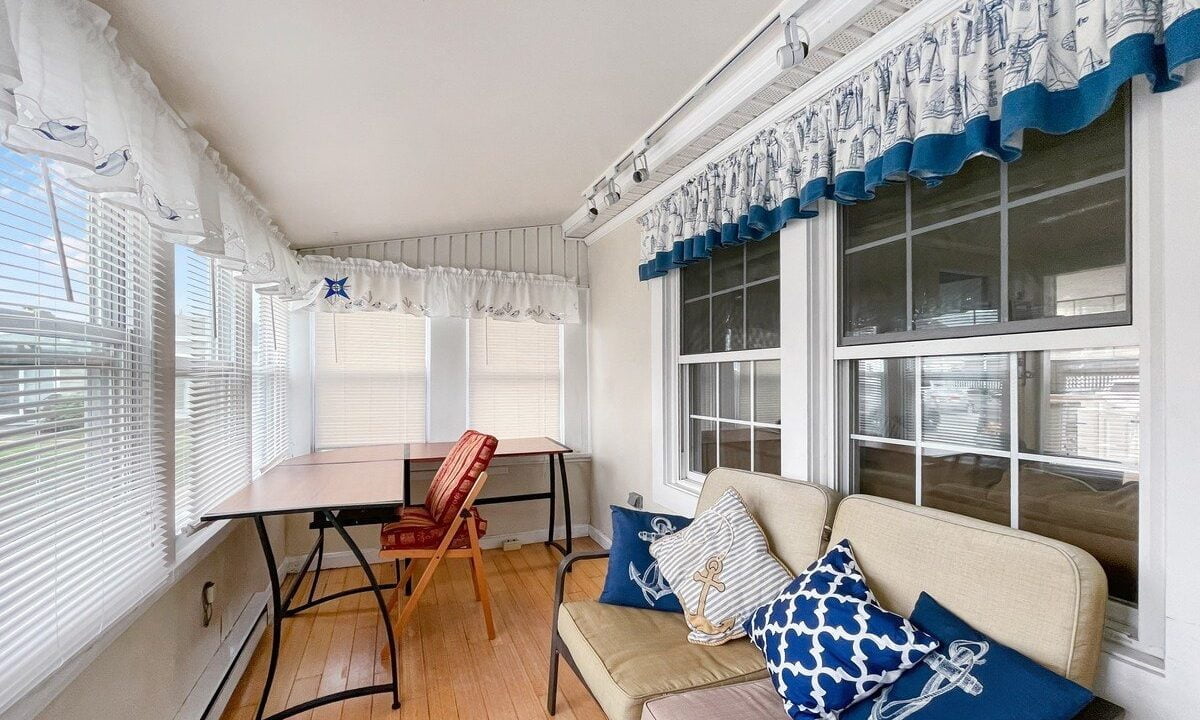Bright sunroom with a beige couch, decorative pillows, a folding table, red chair, and nautical-themed curtains.