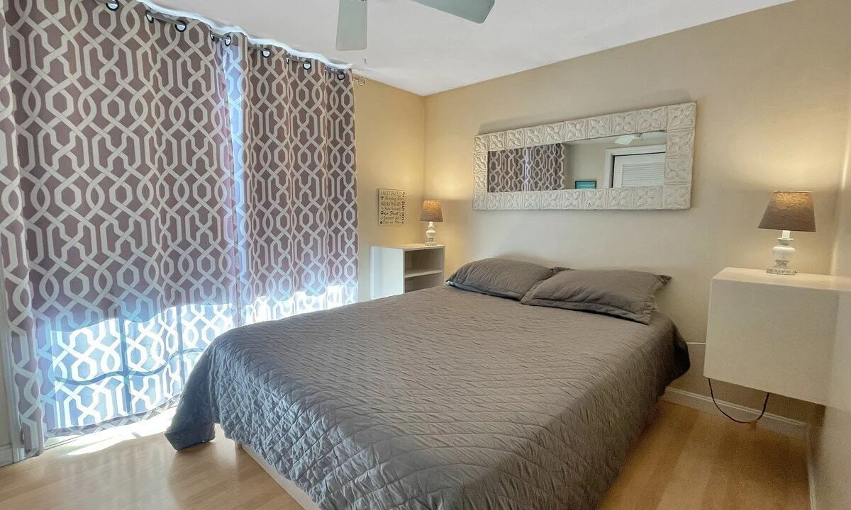 A well-lit bedroom with a large bed covered in a gray duvet, patterned curtains, ceiling fan, mirrored wall decor, and two bedside tables with lamps.