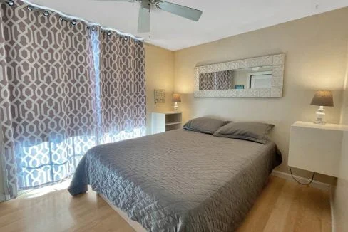 A well-lit bedroom with a large bed covered in a gray duvet, patterned curtains, ceiling fan, mirrored wall decor, and two bedside tables with lamps.