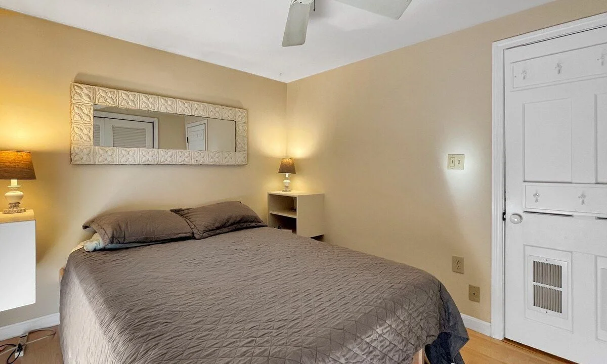 A neat bedroom featuring a large bed with gray bedding, twin nightstands with lamps, a ceiling fan above, and a closed door on the right.