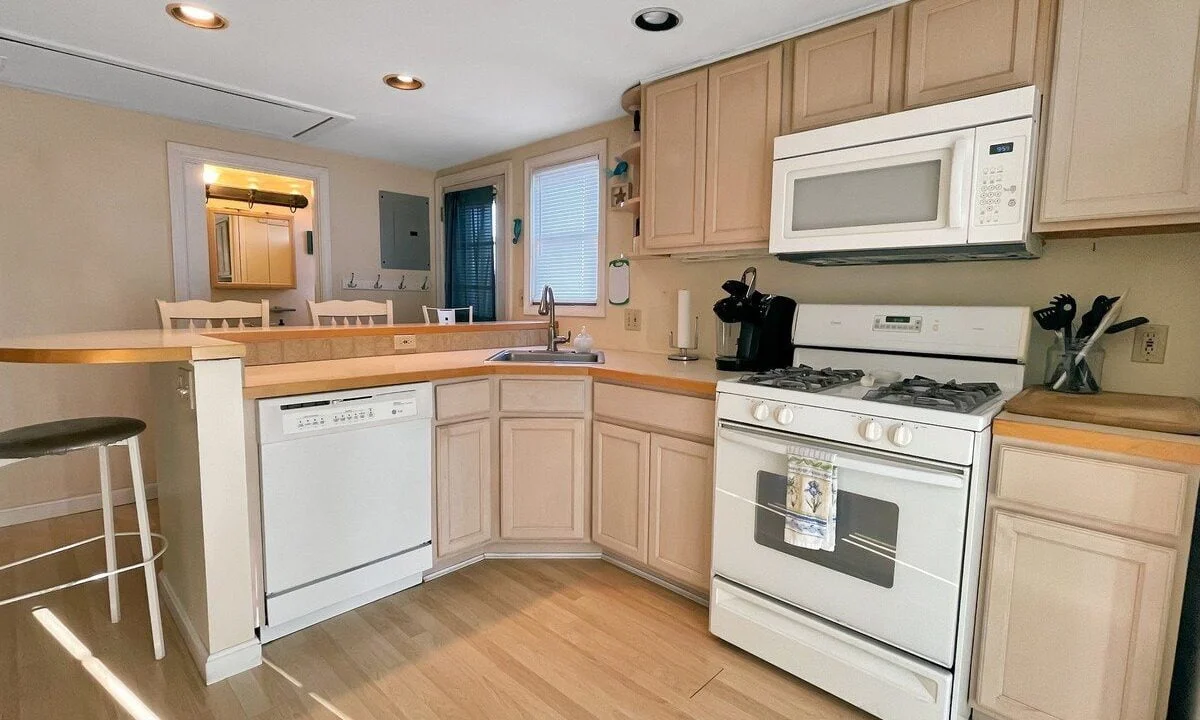 A well-lit kitchen with light wood cabinets, white appliances, and a breakfast bar with a stool.