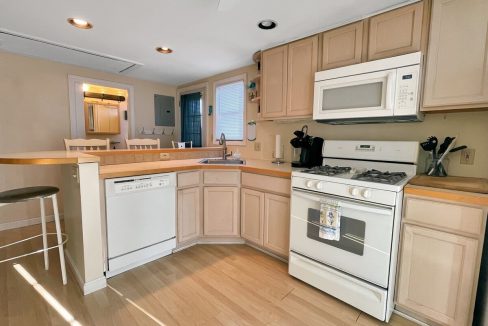 A well-lit kitchen with light wood cabinets, white appliances, and a breakfast bar with a stool.