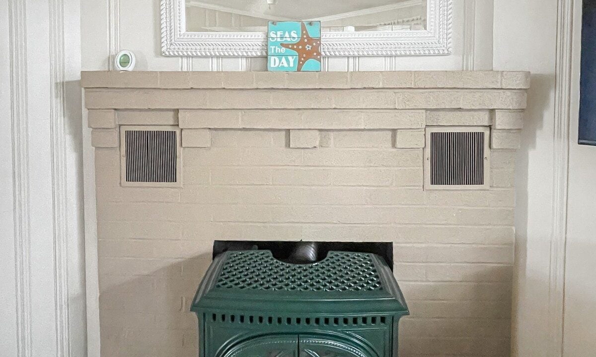 A green vintage-style stove stands in front of a bricked fireplace alcove, flanked by two vents, with a mirror hanging above and a book titled 