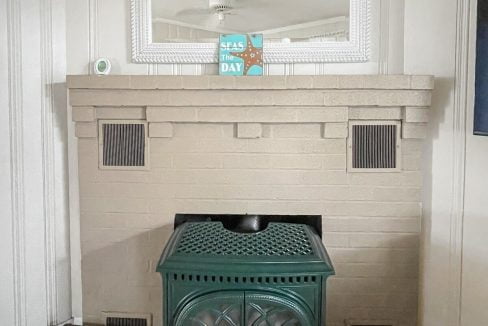 A green vintage-style stove stands in front of a bricked fireplace alcove, flanked by two vents, with a mirror hanging above and a book titled 