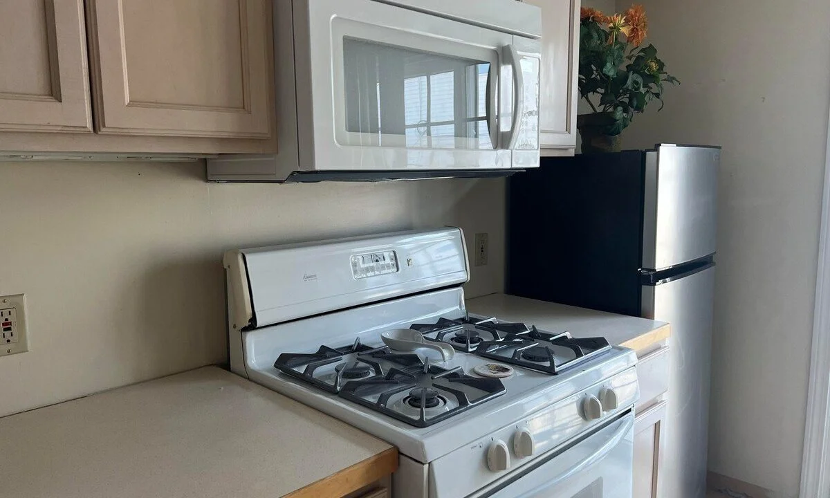 A compact kitchen with a gas stove, overhead microwave, light wood cabinets, a countertop, and a refrigerator placed in the corner.