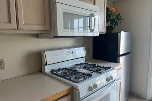 A compact kitchen with a gas stove, overhead microwave, light wood cabinets, a countertop, and a refrigerator placed in the corner.