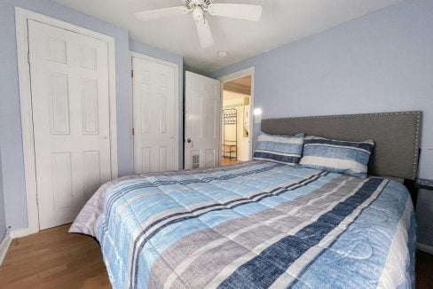 A tidy bedroom featuring a bed with a blue and gray striped comforter, wooden flooring, white doors, and a ceiling fan.