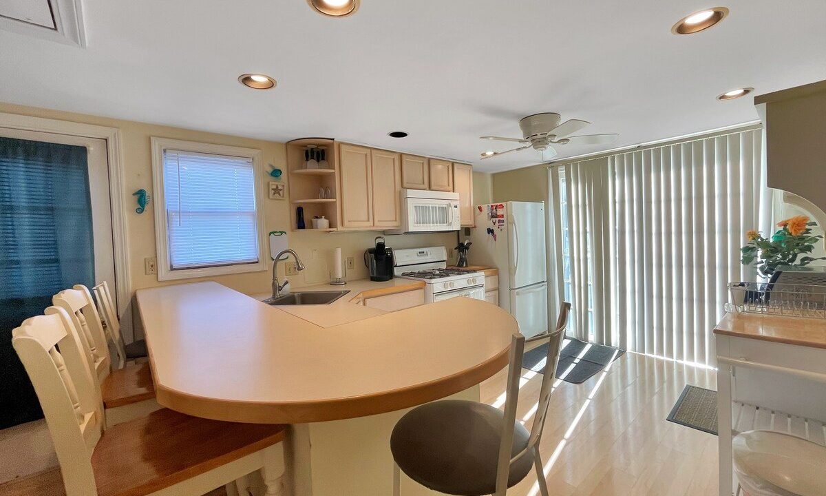 Bright kitchen with beige cabinets, curved breakfast bar with stools, overhead lighting, and an adjacent dining area with a large window covered by vertical blinds.