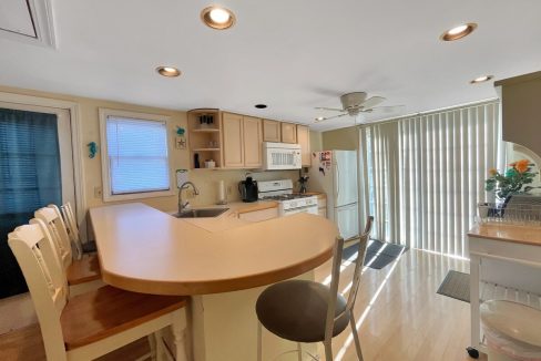 Bright kitchen with beige cabinets, curved breakfast bar with stools, overhead lighting, and an adjacent dining area with a large window covered by vertical blinds.