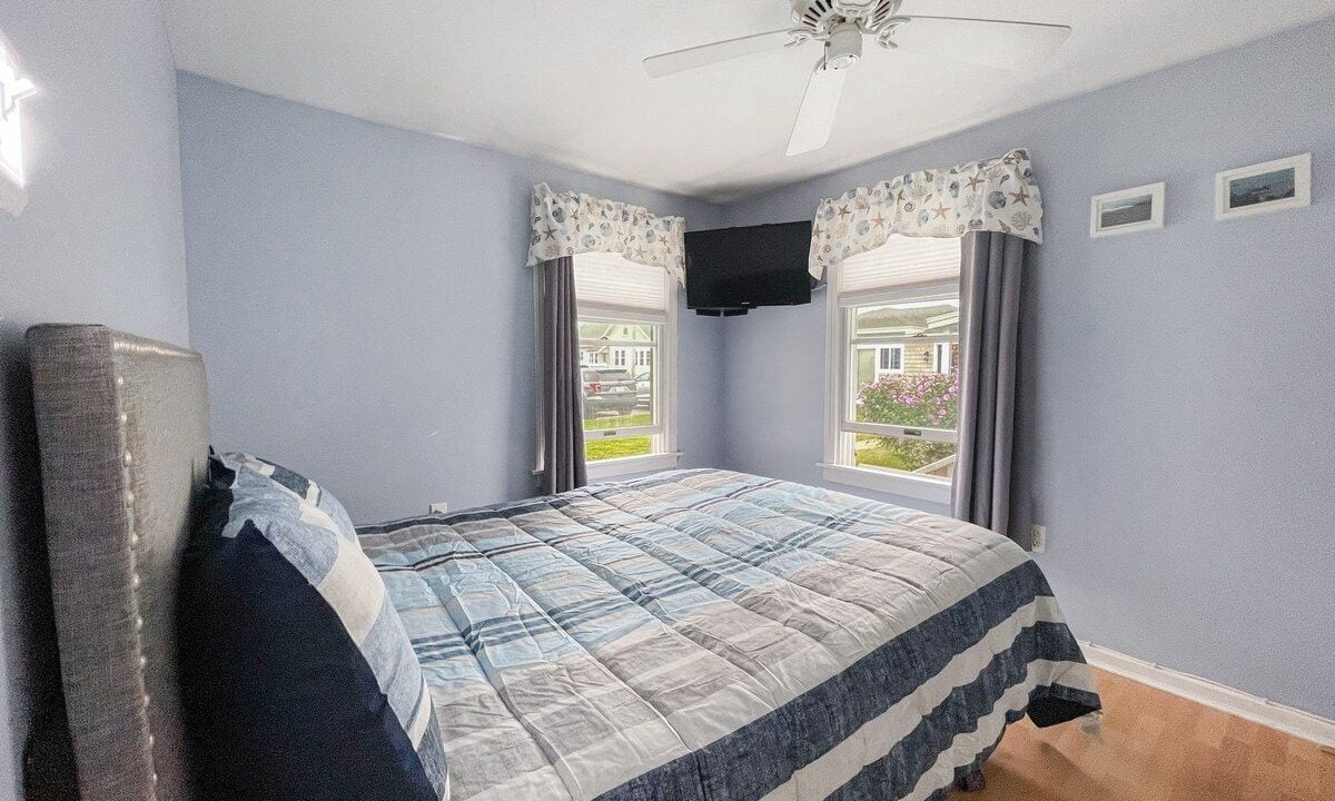 A neat bedroom with light blue walls, a bed with a blue and gray striped comforter, hardwood floor, a ceiling fan, and windows showing a residential view.