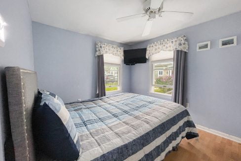 A neat bedroom with light blue walls, a bed with a blue and gray striped comforter, hardwood floor, a ceiling fan, and windows showing a residential view.