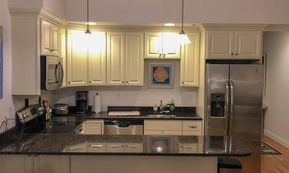 A modern kitchen with wooden flooring, dark granite countertops, white cabinets, a stainless steel refrigerator, microwave, and two pendant lights above a breakfast bar with two stools.