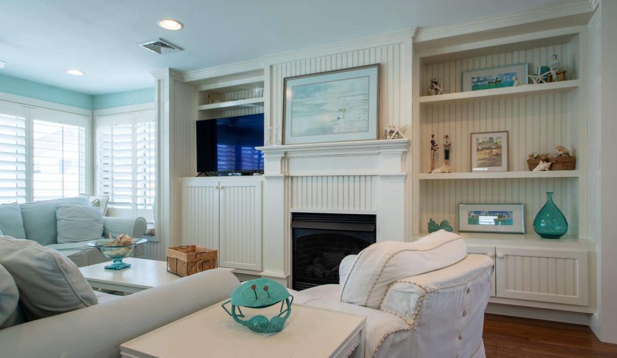 A living room with light blue and white decor features a fireplace with built-in shelving, a blue couch, a white armchair, and beach-themed accessories. A TV is mounted above the fireplace.