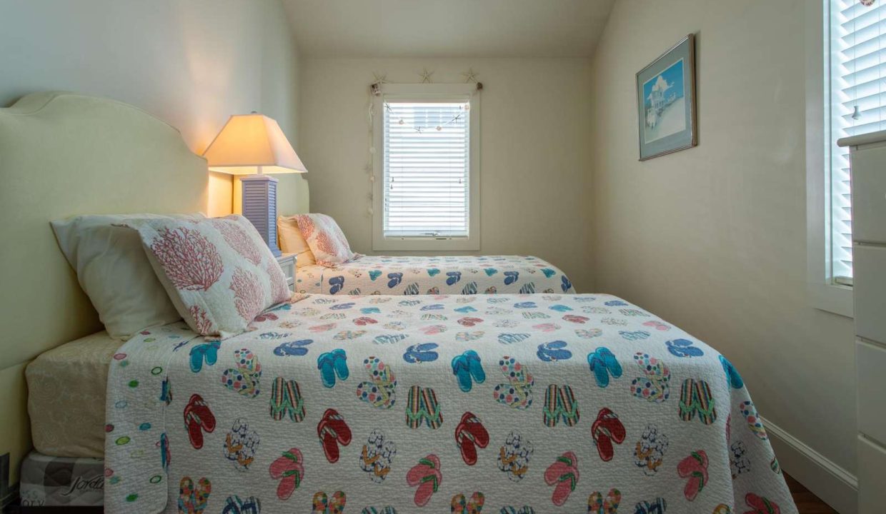 A small bedroom with two single beds, each covered with a quilt featuring colorful flip-flop patterns. A lamp is on a nightstand between the beds, and a window with blinds is in the background.
