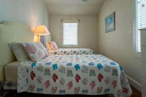 A small bedroom with two single beds, each covered with a quilt featuring colorful flip-flop patterns. A lamp is on a nightstand between the beds, and a window with blinds is in the background.