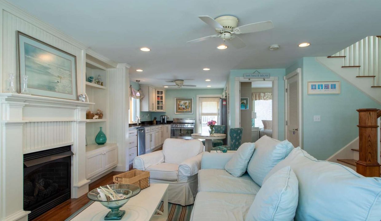 A cozy living room with light blue furniture, a fireplace, and an open layout leading to the kitchen. The space features a ceiling fan, staircase, and various decorative elements.